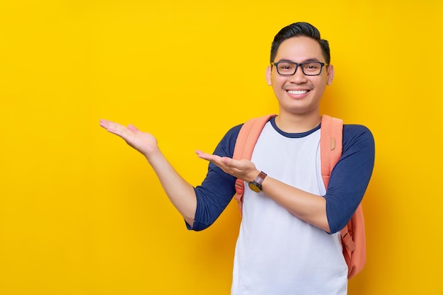 Smiling young asian boy student wearing casual clothes and backpack showing open hand palm with copy space new advertising presentation with hand isolated on yellow background