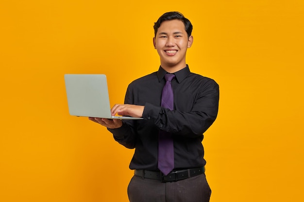 Улыбающийся молодой азиатский бизнесмен, держащий и использующий ноутбук, изолированные на желтом фоне
