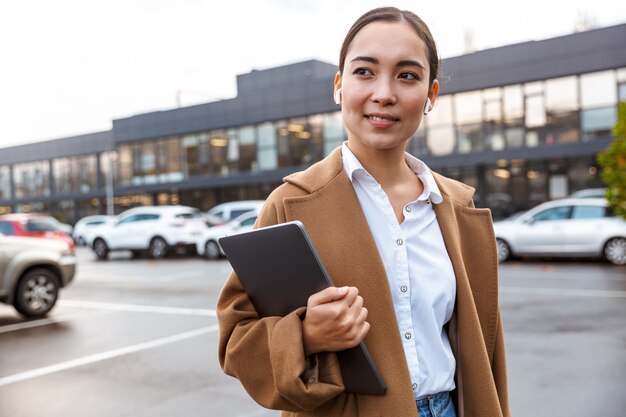 Smiling young asian businessowoman wearing coat walking outdoors in the city, carrying laptop