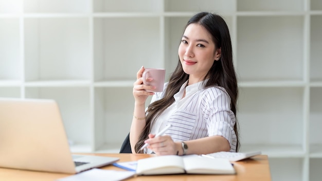 Улыбающаяся молодая азиатская деловая женщина с кружкой кофе и ноутбуком в офисе смотрит в камеру