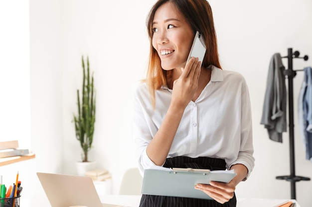 Smiling young asian businesswoman looking through paperwork while talking on mobile phone at the office