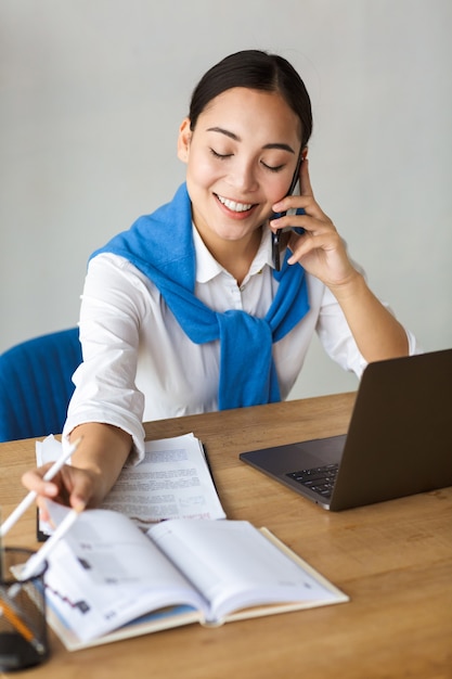 Sorridente giovane imprenditrice asiatica che lavora in ufficio, parla al cellulare mentre lavora al computer portatile