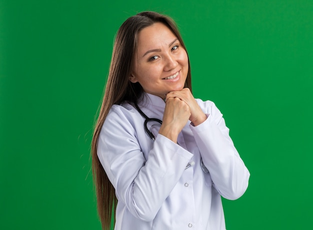 Sorridente giovane dottoressa asiatica che indossa abito medico e stetoscopio tenendo le mani insieme sotto il mento guardando la parte anteriore isolata sulla parete verde con spazio di copia