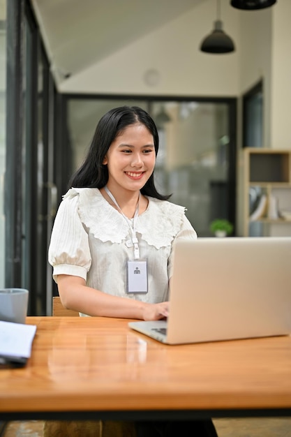 Smiling young Asian female secretary or office worker is working on her task on laptop