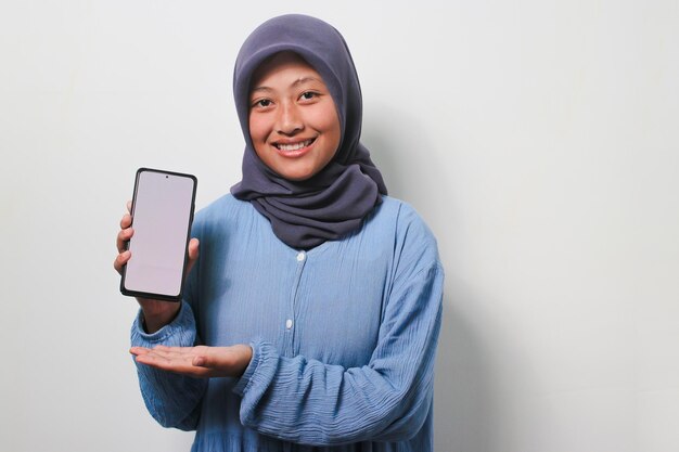 Фото Улыбающаяся молодая азиатка в хиджабе показывает белый пустой экран мобильного телефона с копировальным пространством