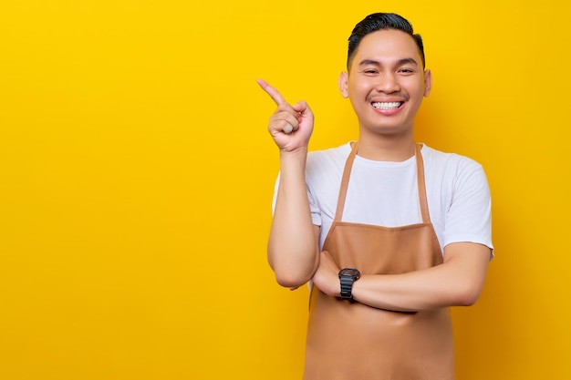 Smiling young Asian man 20s barista barman employee wearing brown apron working in coffee shop pointing finger on advertising area on yellow background Small business startup concept