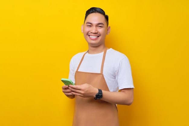 Smiling young Asian man barista barman employee wearing brown apron work in coffee shop holding mobile phone isolated on yellow background Small business startup concept