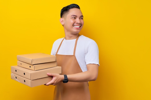 Smiling young Asian man barista barman employee wearing brown apron working in coffee shop holding italian pizza in cardboard and looking aside on yellow background Small business startup concept