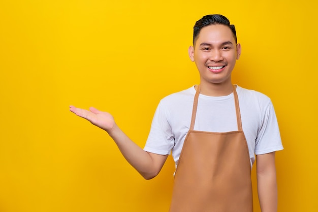 Smiling young asian man barista barman employee wearing brown apron working in coffee shop pointing hand aside isolated on yellow background small business startup concept