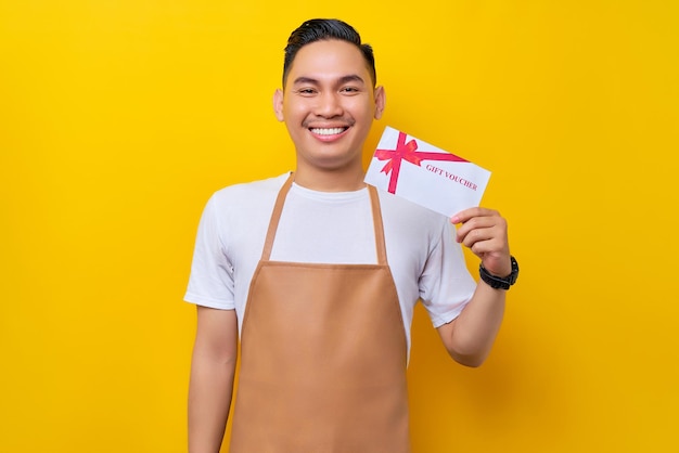 Smiling young Asian man barista barman employee wearing a brown apron working in a coffee shop showing a gift certificate coupon voucher card on yellow background Small business startup concept