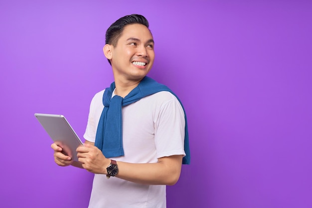 Smiling young Asian man in casual clothes holding digital tablet and looking aside at copy space isolated over purple background