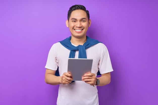 Smiling young asian man in casual clothes holding digital tablet and looking at camera isolated over purple background