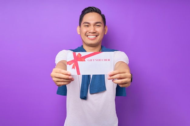 Smiling young asian man in casual clothes holding gift voucher certificate isolated on purple studio background