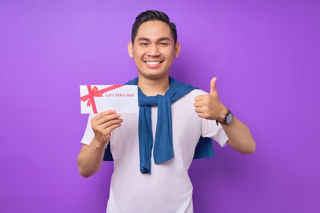 smiling young asian man in casual clothes holding gift voucher certificate and showing thumb up isolated on purple studio background