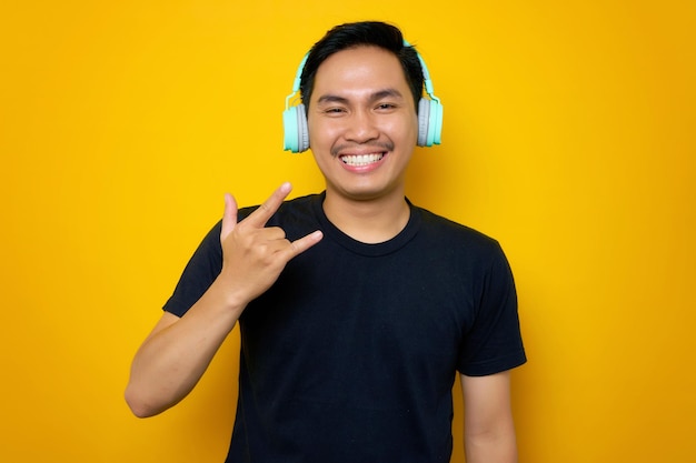 Улыбающийся молодой азиат в повседневной футболке слушает музыку в наушниках, символ металлического рока, изолированный на желтом фоне.