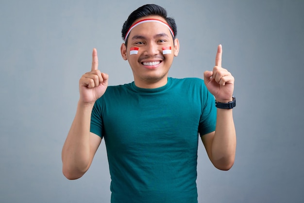 灰色の背景に分離されたスペースをコピーするために指を指すカジュアルなTシャツで若いアジア人の笑顔インドネシア独立記念日のお祝いのコンセプト