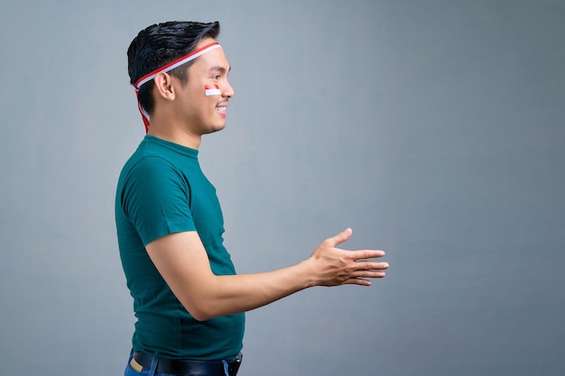 Foto sorridente giovane asiatico in maglietta casual allungando la mano per la stretta di mano isolata su sfondo grigio concetto di celebrazione del giorno dell'indipendenza dell'indonesia