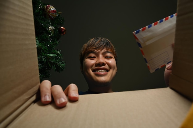 Улыбающийся молодой азиатский мужчина держит поздравительную открытку, заглядывая внутрь картонной коробки.