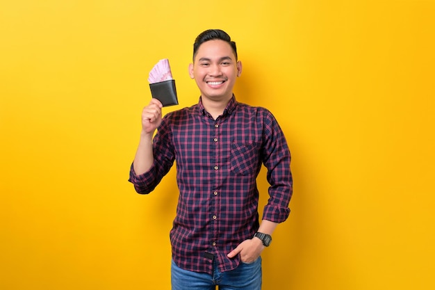 Smiling young Asian man holding wallet full of money banknotes isolated over yellow background Profit and wealth concept