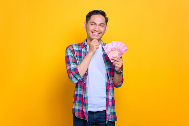 Smiling young Asian man in plaid shirt holding money banknotes and looking at camera isolated on yellow background