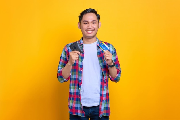 Smiling young Asian man in plaid shirt holding wallet and showing credit card isolated on yellow background