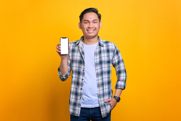 Smiling young Asian man in plaid shirt showing mobile phone blank screen recommending app isolated on yellow background