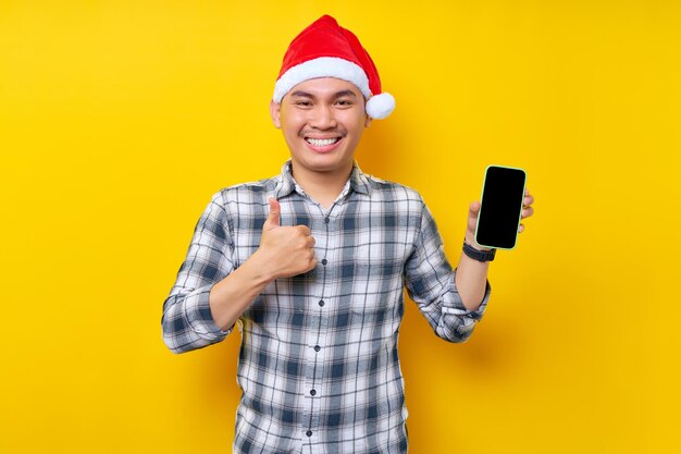 Smiling young asian man in plaid shirt wearing christmas hat\
holding mobile phone with blank screen showing thumb up on yellow\
background christmas and happy new year 2023 holiday concept