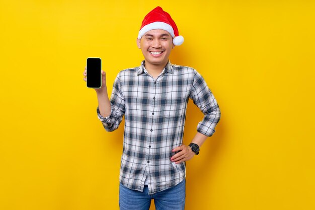 Smiling young asian man in plaid shirt wearing christmas hat\
holding mobile phone with blank screen workspace area on yellow\
background christmas and happy new year 2023 holiday concept