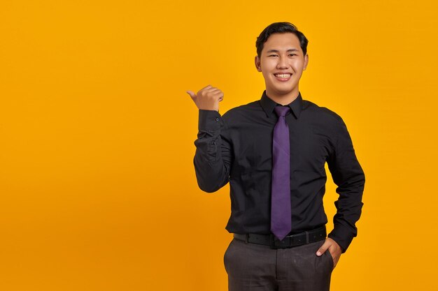 Smiling young Asian man pointing fingers at copy space on yellow background