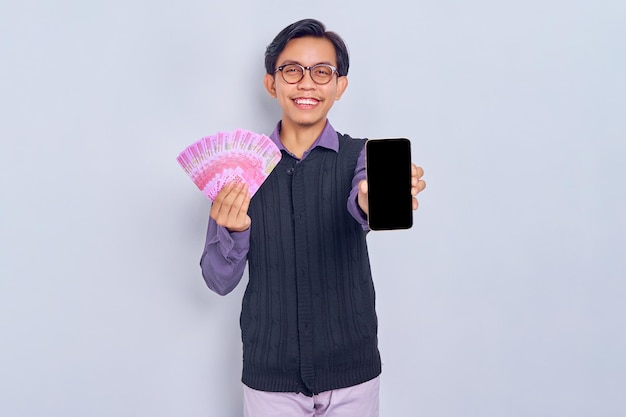 Smiling young Asian man in purple shirt clothes showing blank screen mobile phone and holding cash money in rupiah banknotes isolated on white background People lifestyle concept