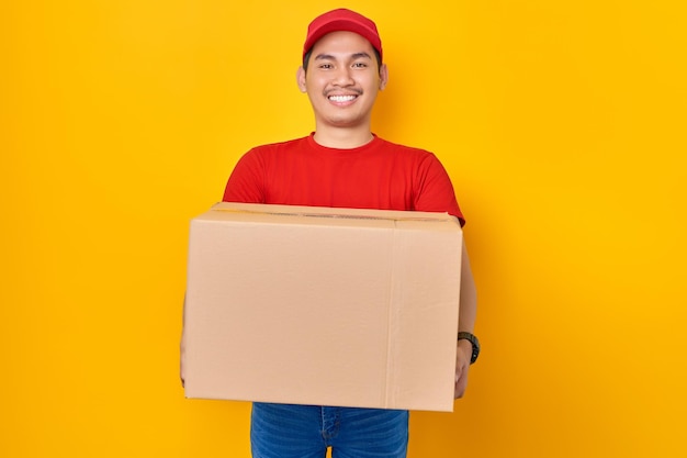 Улыбающийся молодой азиат в красной кепке, служащий в форме футболки, работает курьером-дилером, раздавая коробку с посылкой клиенту, изолированному на желтом фоне. Профессиональная концепция службы доставки