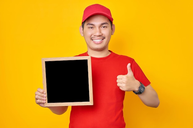 赤い帽子の t シャツの制服従業員で笑顔の若いアジア人男性黄色の背景に親指アップ ジェスチャーを示す空白の黒板を保持しているディーラー宅配便として働くプロフェッショナル配信サービス コンセプト