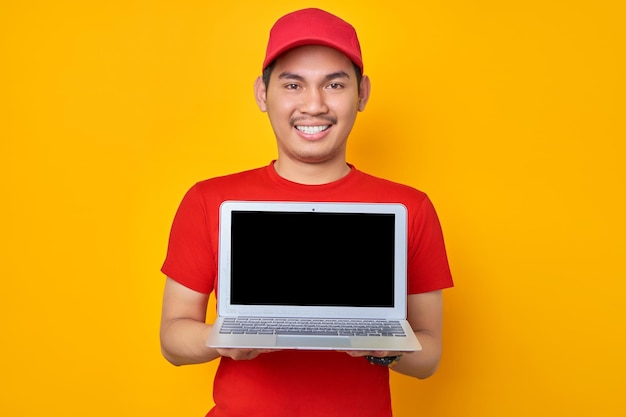 빨간 모자 티셔츠를 입은 웃고 있는 젊은 아시아 남자 직원은 노란색 배경 전문 배달 서비스 개념에 격리된 빈 화면 노트북 컴퓨터를 보여주는 딜러 택배로 일합니다.