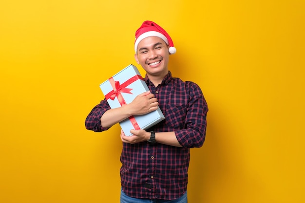 Smiling young Asian man in Santa hat holding gift box with ribbon on yellow studio background Happy New Year 2023 celebration merry holiday concept