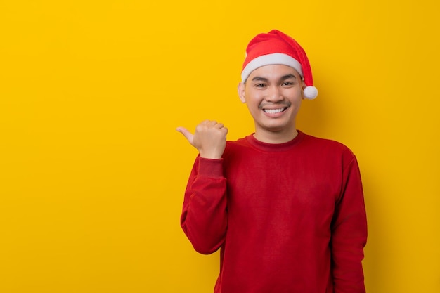 Smiling young Asian man in Santa hat pointing thumb aside at blank space for text on yellow studio background celebration Christmas holiday and New Year concept