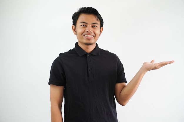 Photo smiling young asian man showing a copy space on a palm and holding another hand on waist