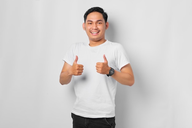 Smiling young Asian man showing thumbs up gesture Approving expression looking at the camera isolated on white background