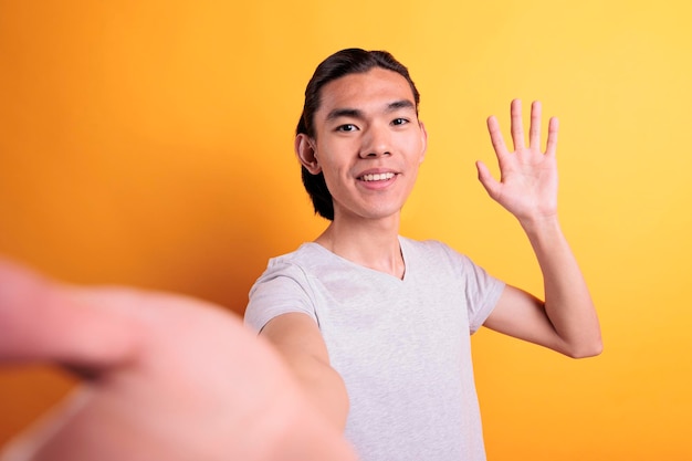 Smiling young asian man talking on video call, waving hello first person view. Mobile phone online communication pov, teleconference conversation, webcam chatting, internet meeting fpv