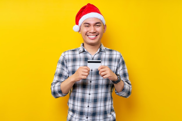 Smiling young asian man wearing christmas hat holding credit\
card on yellow background celebration christmas holiday and new\
year concept