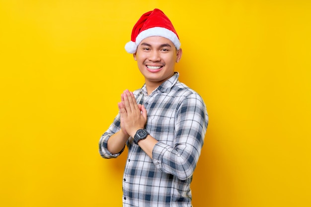 Smiling young Asian man wearing a plaid shirt in a Christmas hat gives greeting hands with a big smile on his face on yellow background celebration Christmas holiday and New Year concept