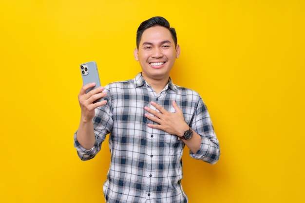 Smiling young asian man wearing a white checkered shirt holding a smartphone and emotionally reacting to online news isolated on yellow background people lifestyle concept