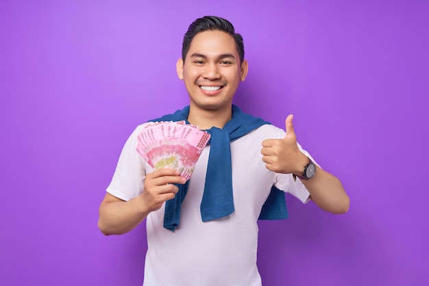 Smiling young asian man wearing white tshirt holding cash money in rupiah banknotes and gesturing thumbs up isolated over purple background people lifestyle concept