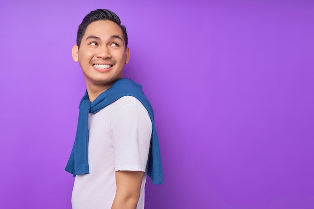 Улыбающийся молодой азиат в белой футболке смотрит в сторону на копировальное пространство, изолированное на фиолетовом фоне, концепция образа жизни людей
