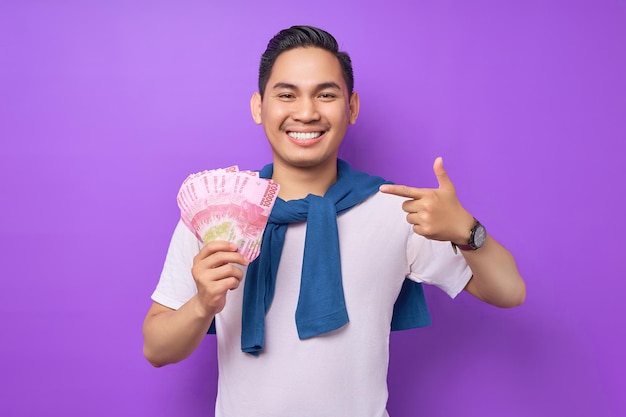 Smiling young Asian man wearing white tshirt pointing fingers at cash money in rupiah banknotes isolated over purple background people lifestyle concept