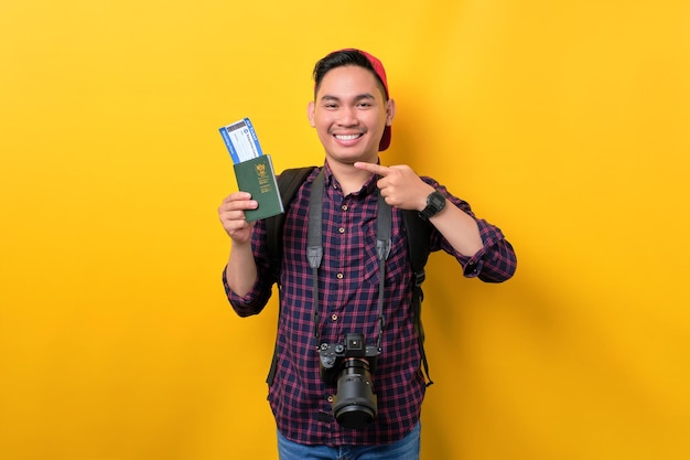 Smiling young Asian man with backpack pointing fingers at passport and flight tickets isolated over yellow background Tourism and holiday trip concept