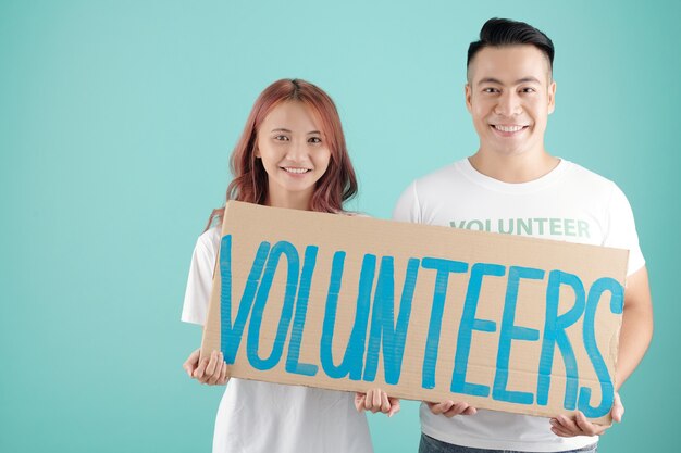 Photo smiling young asian man and woman holding big sign with volunteers inscription