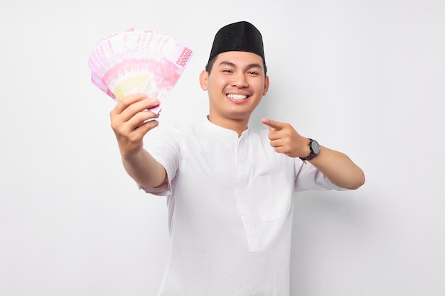 흰색 배경에 고립 된 현금 돈 루피아 지폐에 손가락을 가리키는 젊은 아시아 무슬림 남자 웃고 사람들 종교 이슬람 라이프 스타일 개념