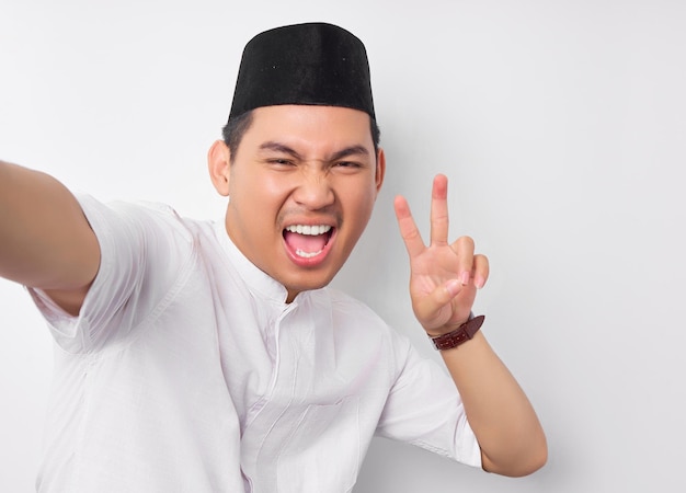 Smiling young Asian Muslim man taking selfie on smartphone showing victory sign isolated on white background Ramadan and eid Mubarak concept