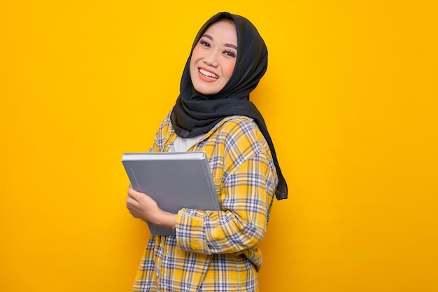 Smiling young asian muslim student holding book looking at\
camera isolated on yellow background back to school in high school\
college concept