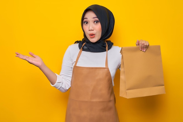 Smiling young Asian Muslim woman barista in white tshirt apron work in coffee shop hold blank paper takeaway bag mock up and point hands arms aside on workspace area isolated on yellow background